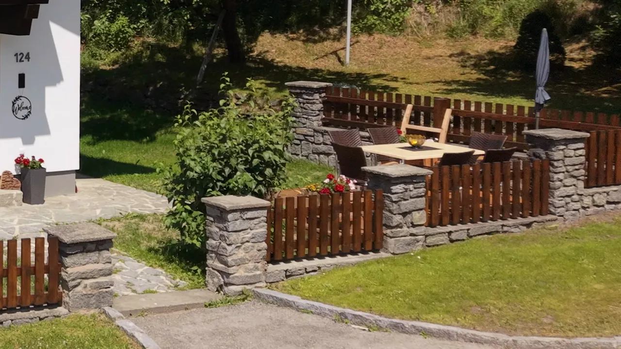Blick in den Garten: grüne Wiese, Sträucher und die eingezäunte Sitzecke mit Sonnenschirm direkt neben dem Haus