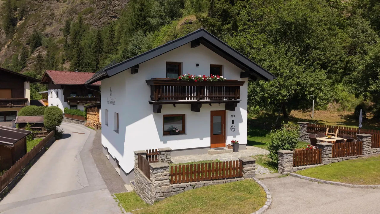 Das Ferienhäusl aus der Nähe: weiße Fassade mit handgemaltem Schriftzug, Balkon voller Geranien, rechts die Terrasse hinter der Steinmauer
