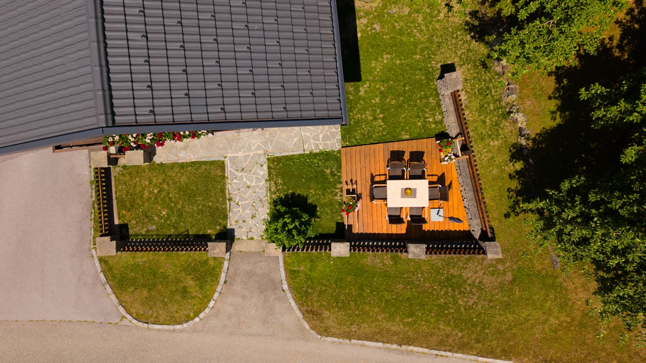 Die Holzterrasse aus der Nähe: großer Tisch mit Stühlen, daneben Blumen und Rasen