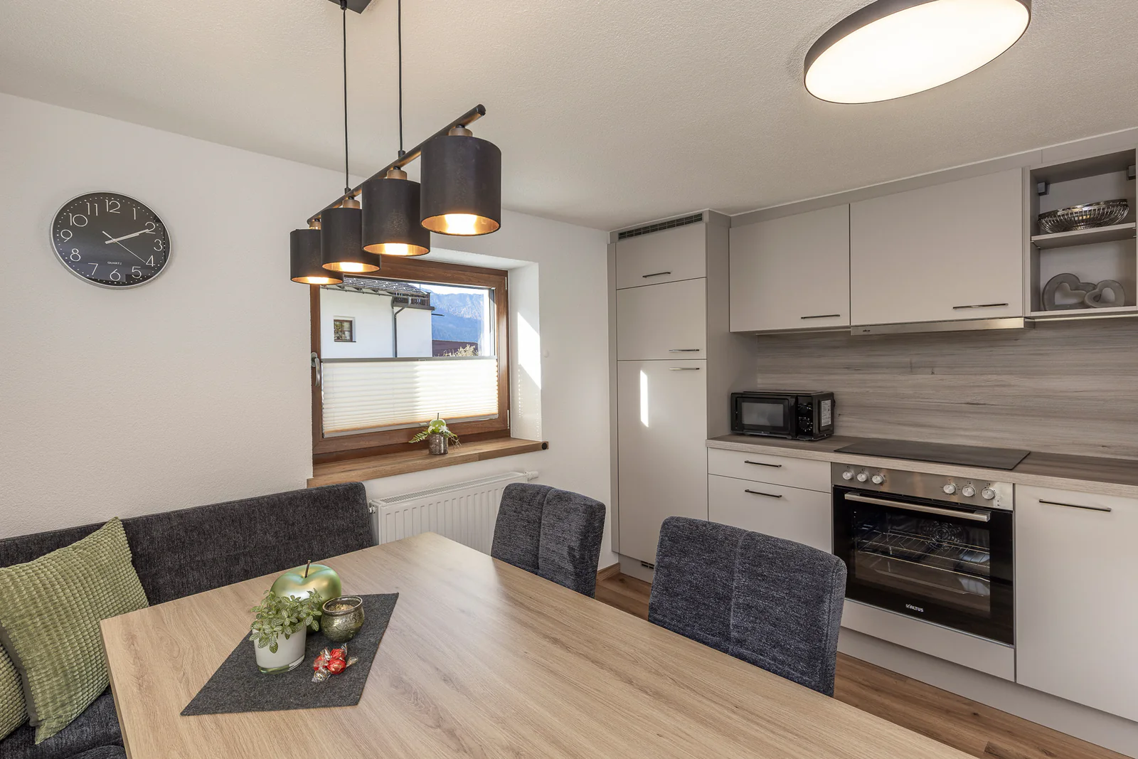 Die Wohnküche: Esstisch mit Eckbank und salbeigrünen Kissen, dahinter die Küchenzeile mit Backofen